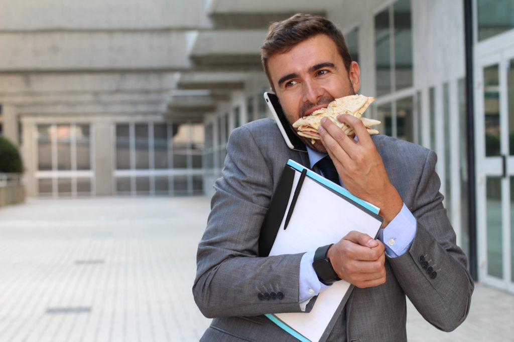 Multitasking businessman holding folders and phone to his shoulder while walking and eating a sandwich