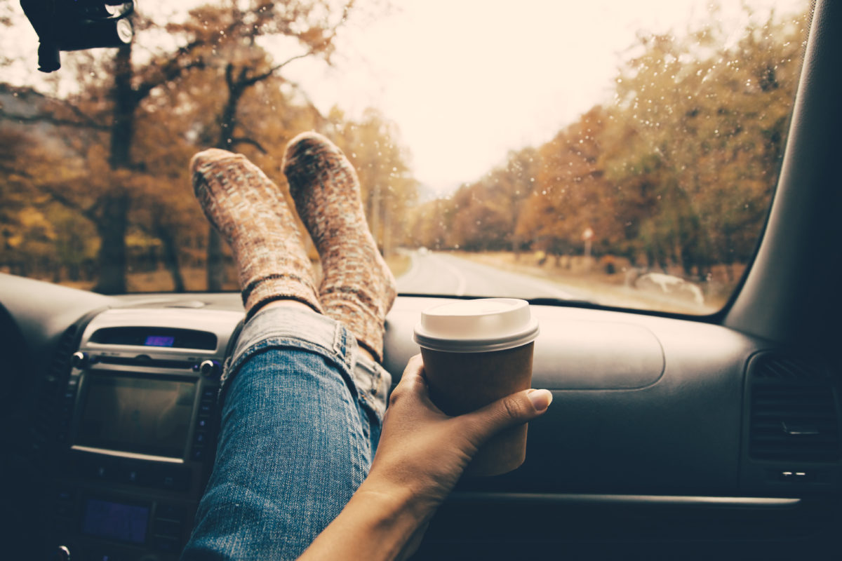 Socked feet on dashboard of moving car with hand holding coffee